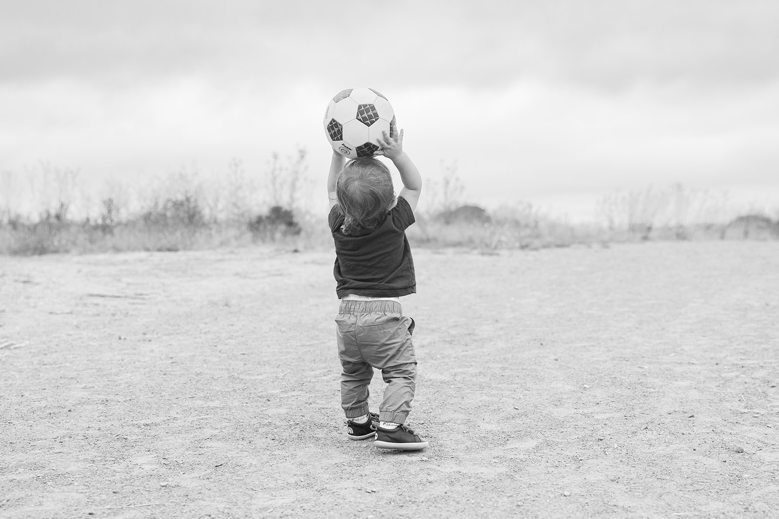 Black and white photo of a toddler boy holding a soccer ball above his head