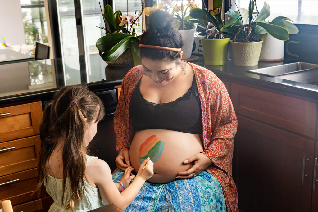 Pregnant mother letting her daughter paint her belly during an at home maternity session in the Bay Area