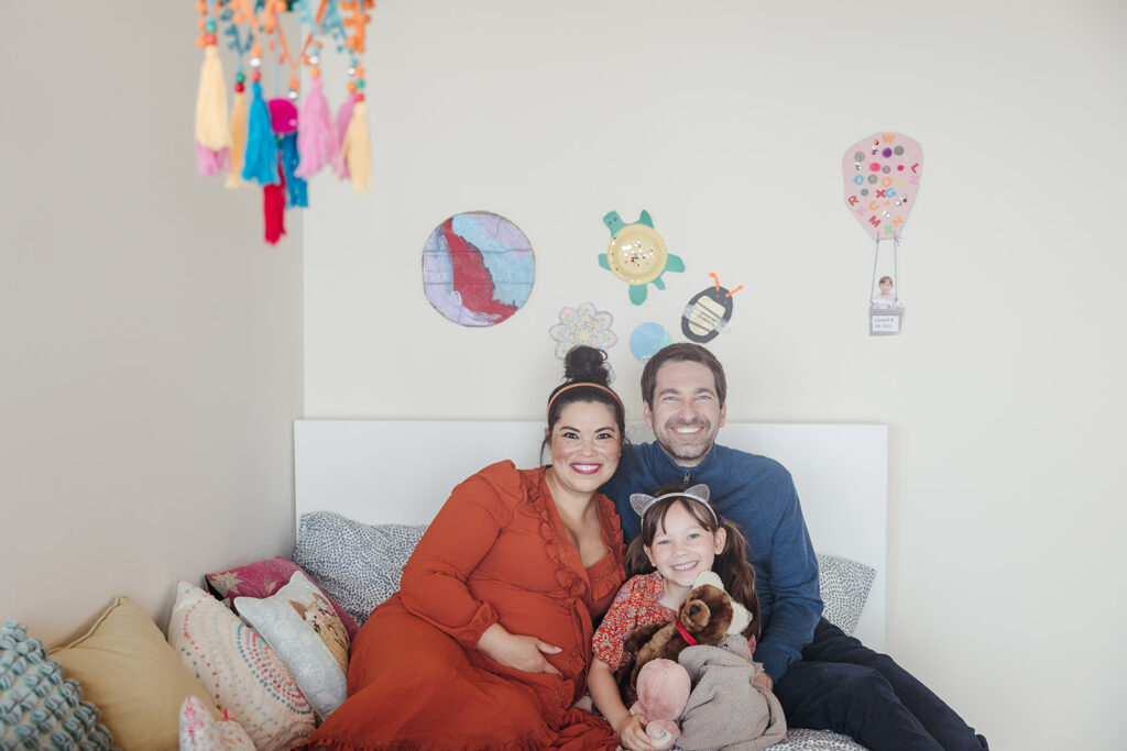 Pregnant mother, father, and daughter sitting on a bed during at home maternity photoshoot