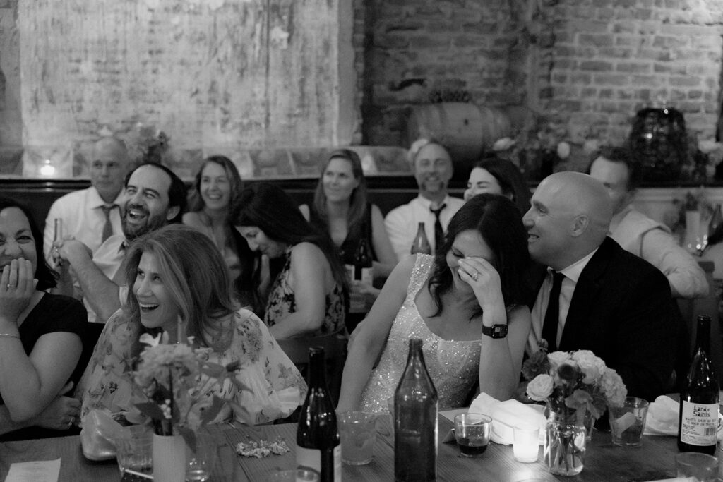 Black and white candid wedding reception photo of a bride and groom laughing during speeches