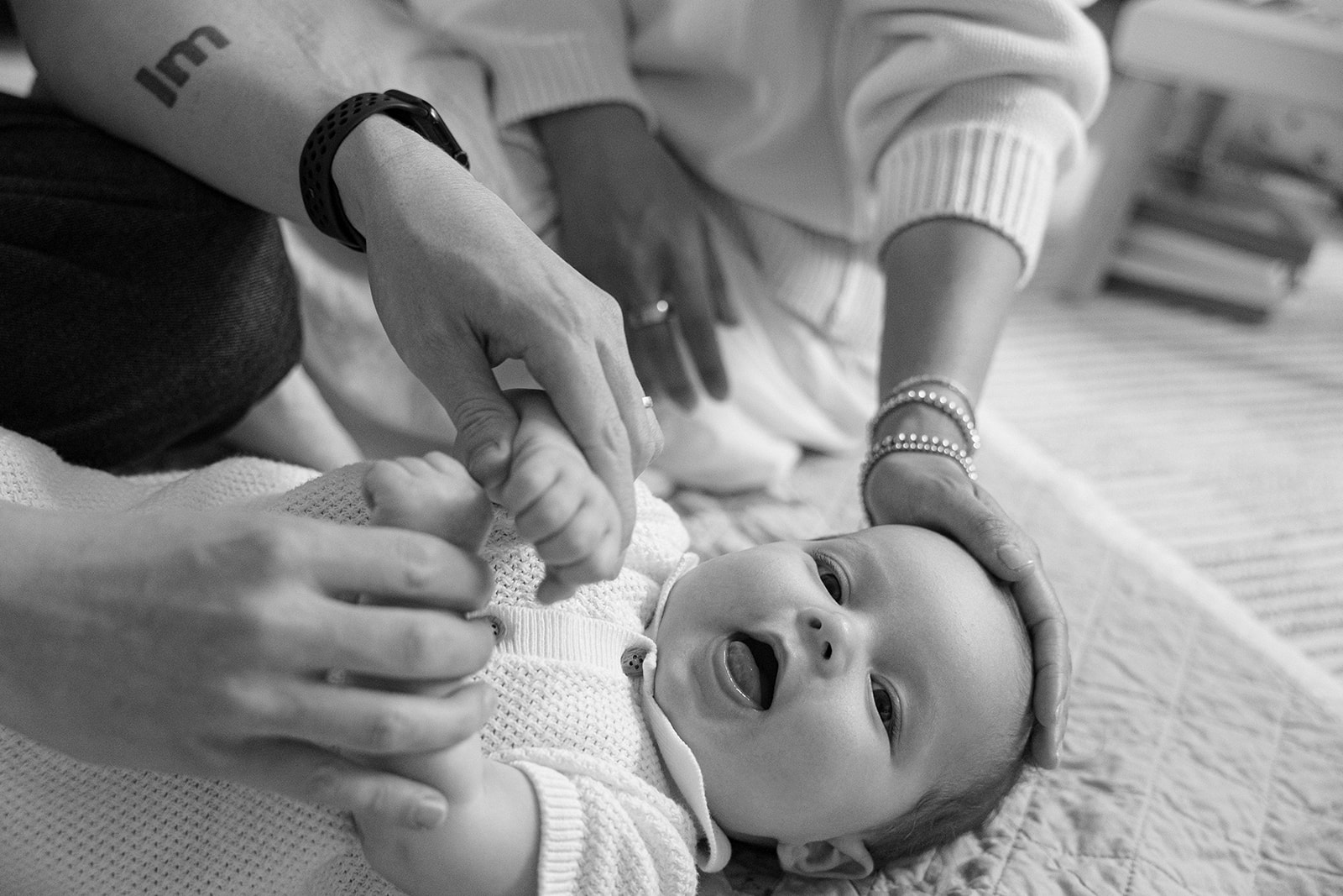 Black and white portrait of a baby laying on the floor with his parents nearby. 