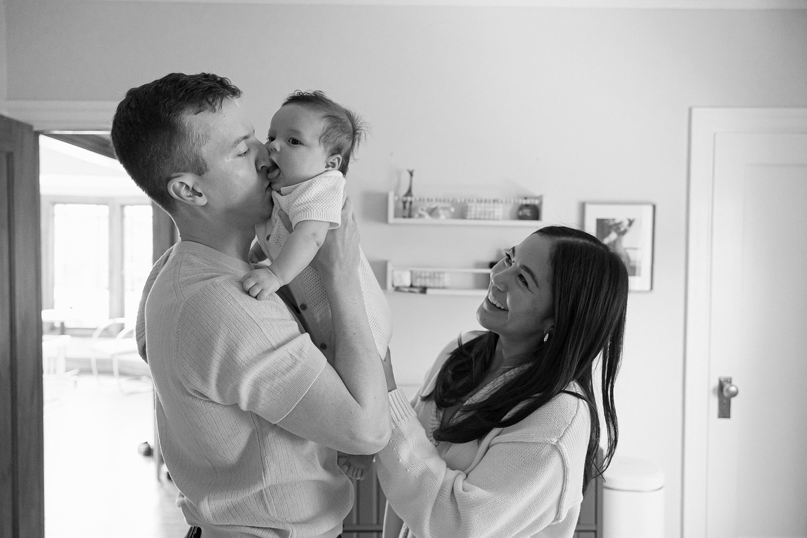 Black and white photo of a baby trying to eat his dads nose with his mother smiling nearby. 