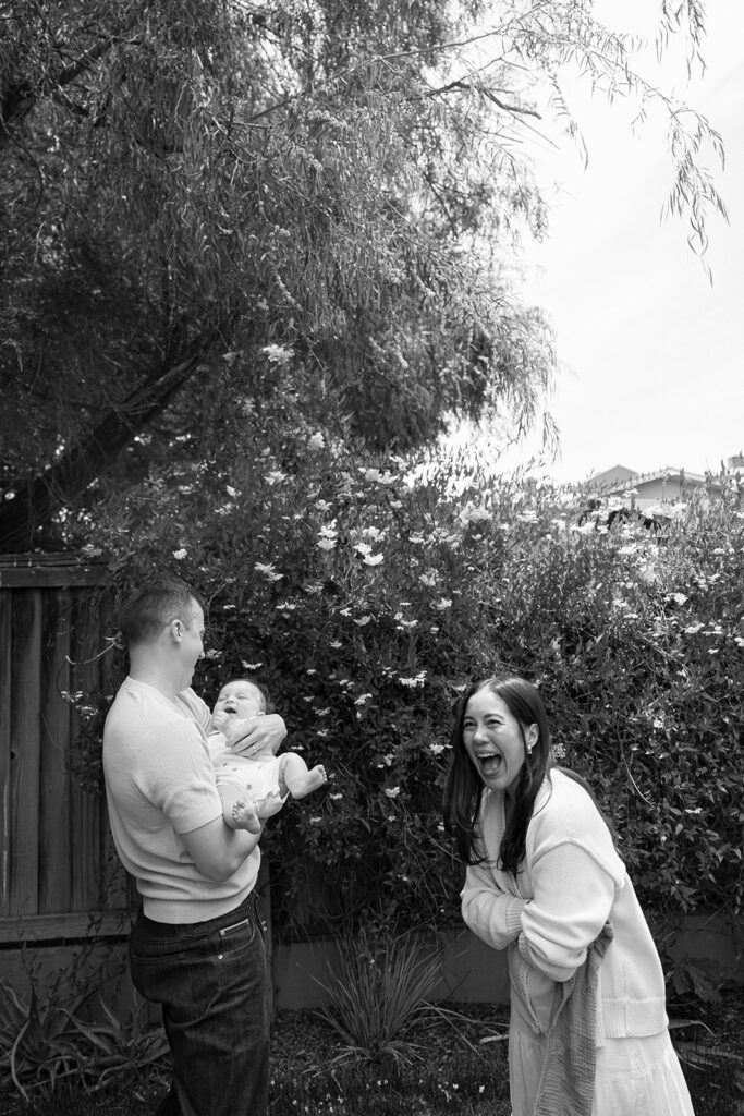 Candid black and white photo of a father holding his small baby with the mother laughing nearby. 