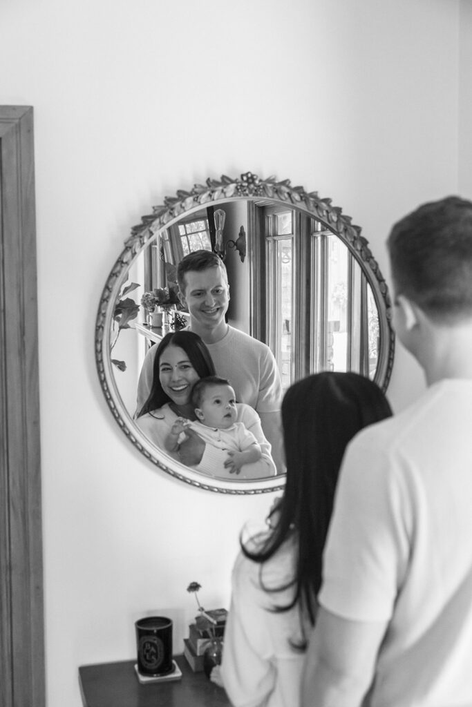 Black and white photo of a mother, father, and their small baby smiling in a mirror during their at-home family photos in Oakland, California. 