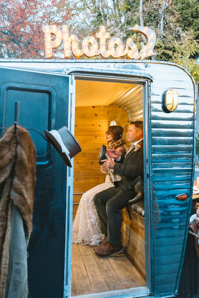 Bride and groom sitting inside photo booth trailer with glowing sign during golden hour at Miners Foundry wedding in Nevada City, California.