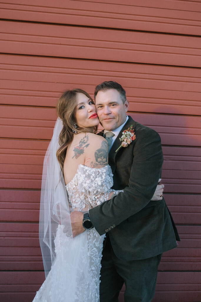 Bride and groom embracing in front of red textured wall during golden hour portraits at Miners Foundry wedding in Nevada City, California.