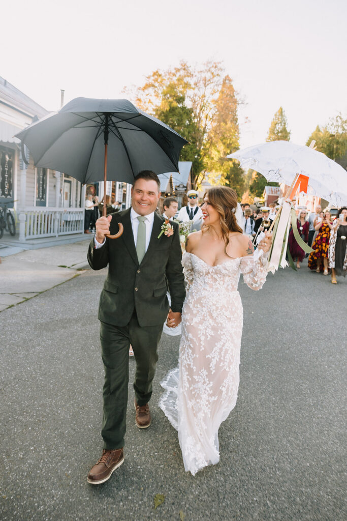 Bride and groom walking hand in hand under umbrellas during marching band processional at Miners Foundry wedding in Nevada City, California.
