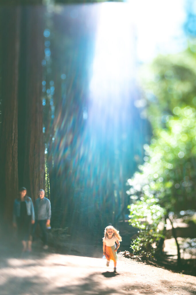 Little girl running through sunlit redwoods during playful Marin maternity session.