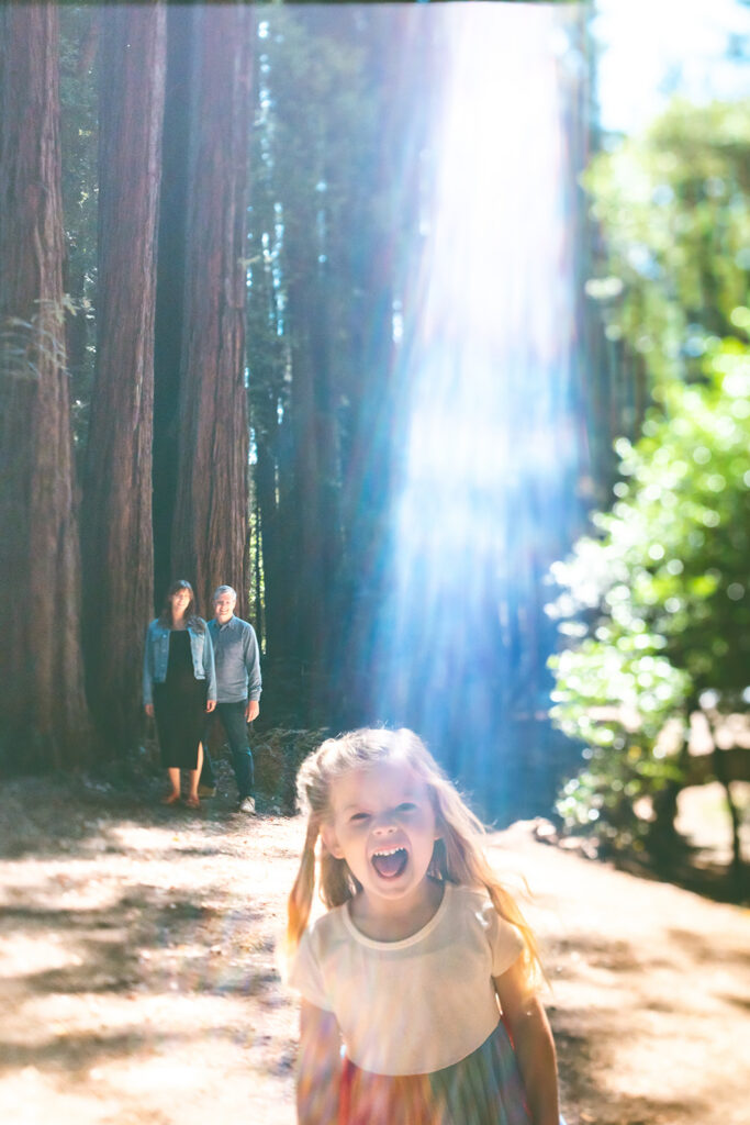 Little girl running through sunlit redwoods during playful Marin maternity session.