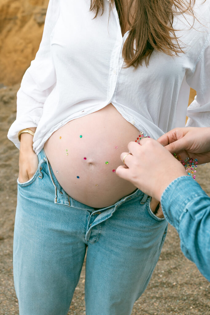 Hands placing colorful jewels on pregnant belly during creative maternity photos at Rodeo Beach.