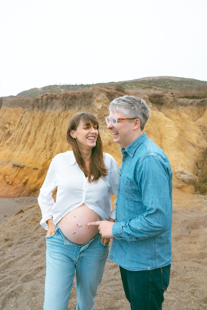 Expectant parents laughing together on coastal cliffs during Rodeo Beach maternity photos.