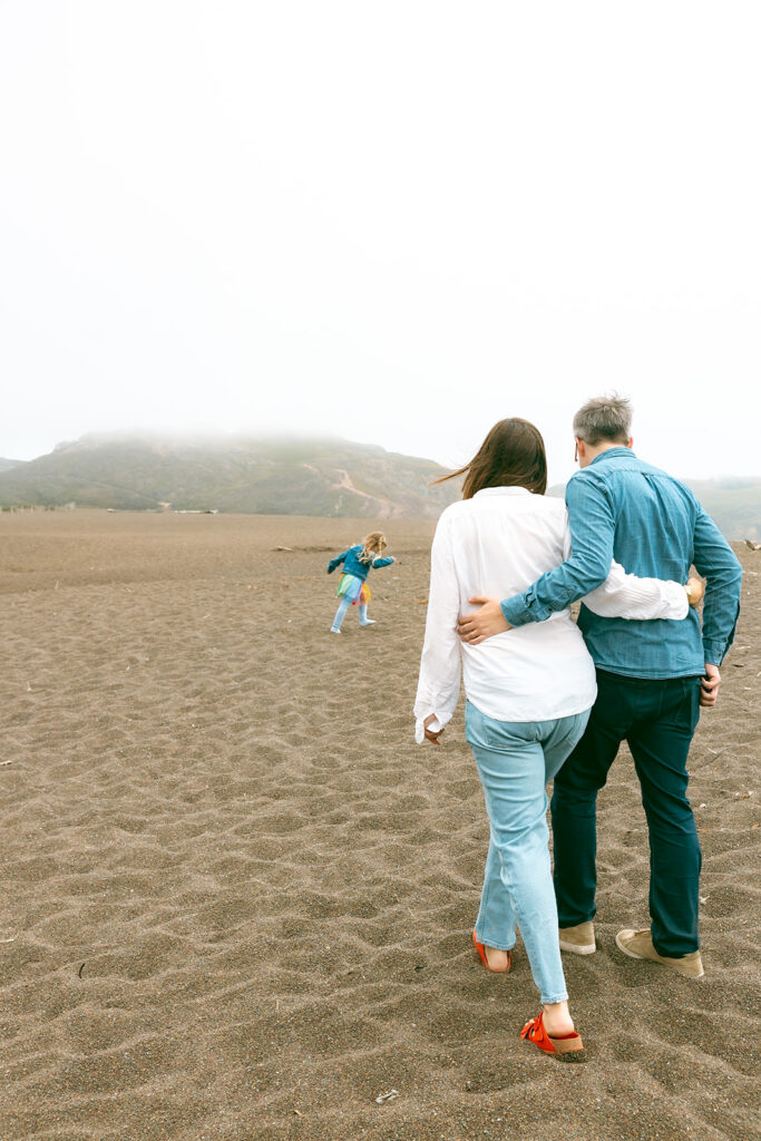 Family walking along Rodeo Beach with older sibling running ahead during maternity session.