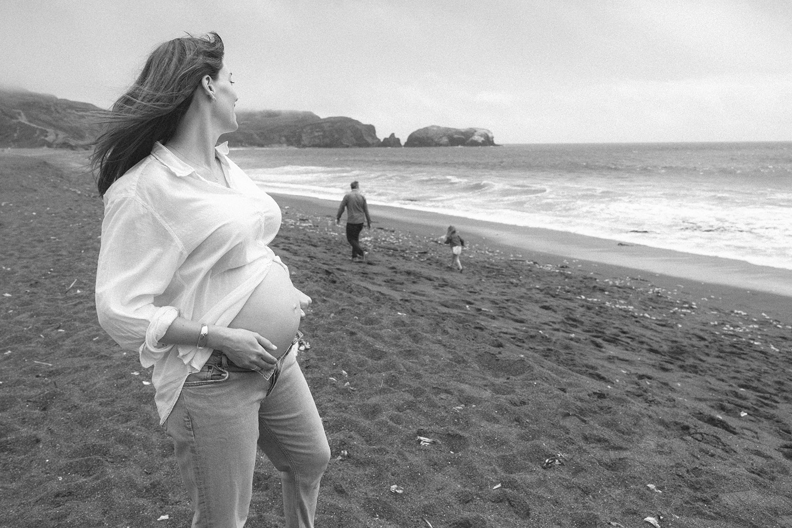 Black and white photo of a mother watching her husband and daughter run along Rodeo Beach during their outdoor maternity photos. 