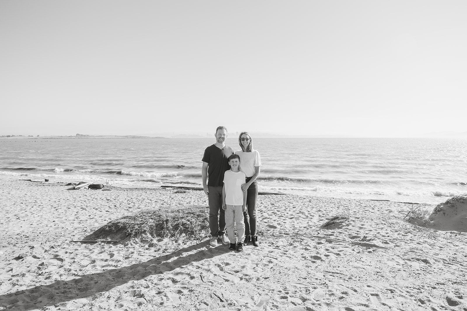 Black and white wide shot of a family of three posing on the beach at Albany Bulb. 