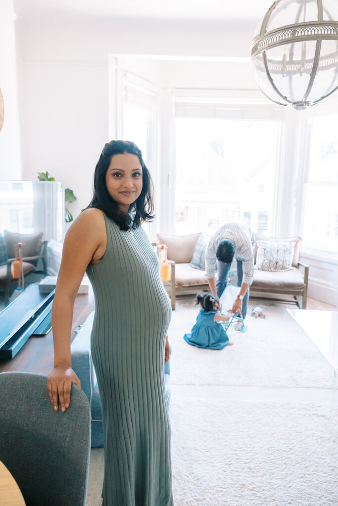 Pregnant mother posing for a photo with her husband and daughter behind her in the living room for their at-home family and San Francisco maternity photos.