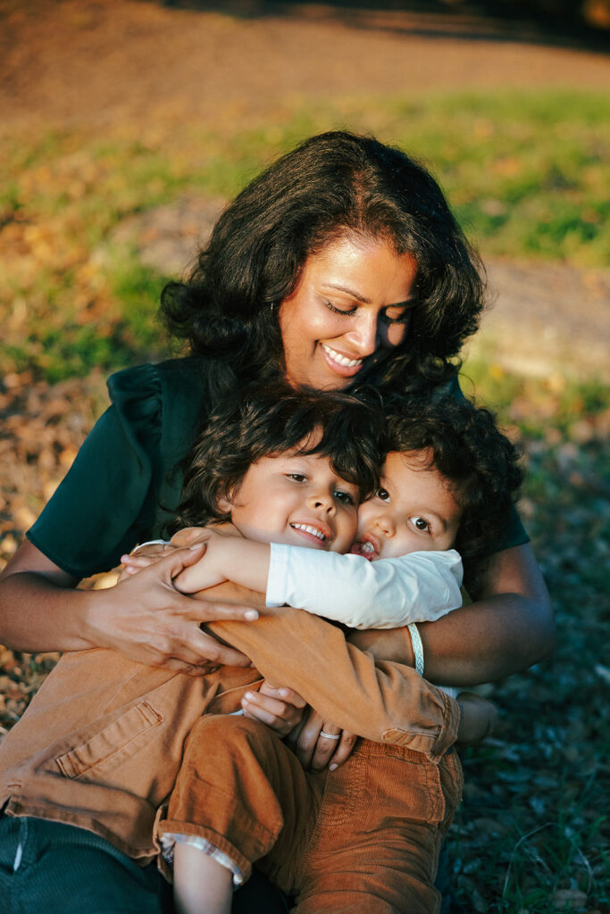 Golden hour photo of a mother smiling and holding her two young son in her arms.