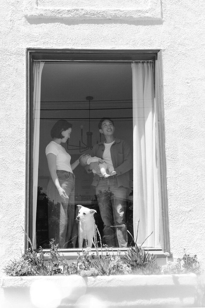 Black and white photo of a couple looking out a window while holding their newborn baby.