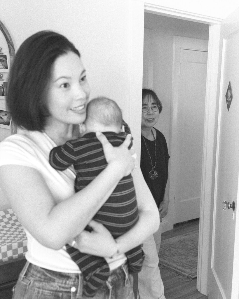 Black and white photo of a mother holding her baby while the grandmother stands in the doorway.