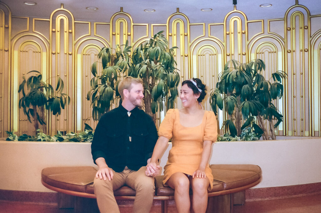 Film engagement photo of a couple sitting together in front of gold details and greenery.