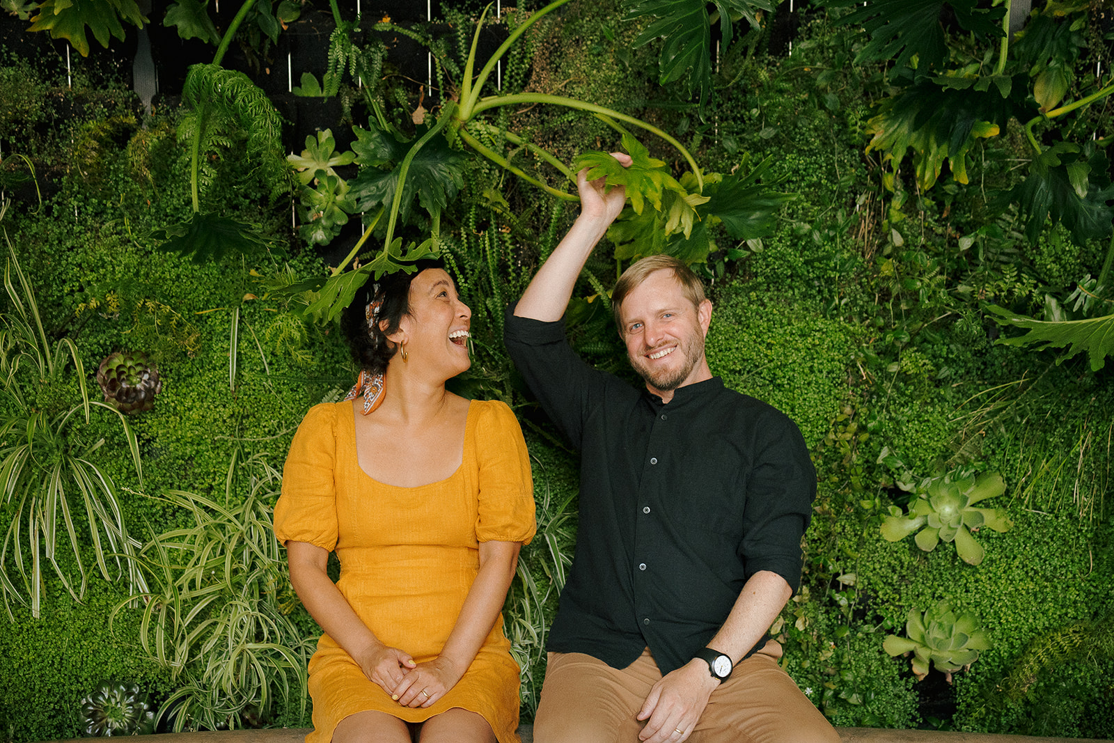 Playful Bay Area engagement photo of a couple laughing in front of a green plant wall.
