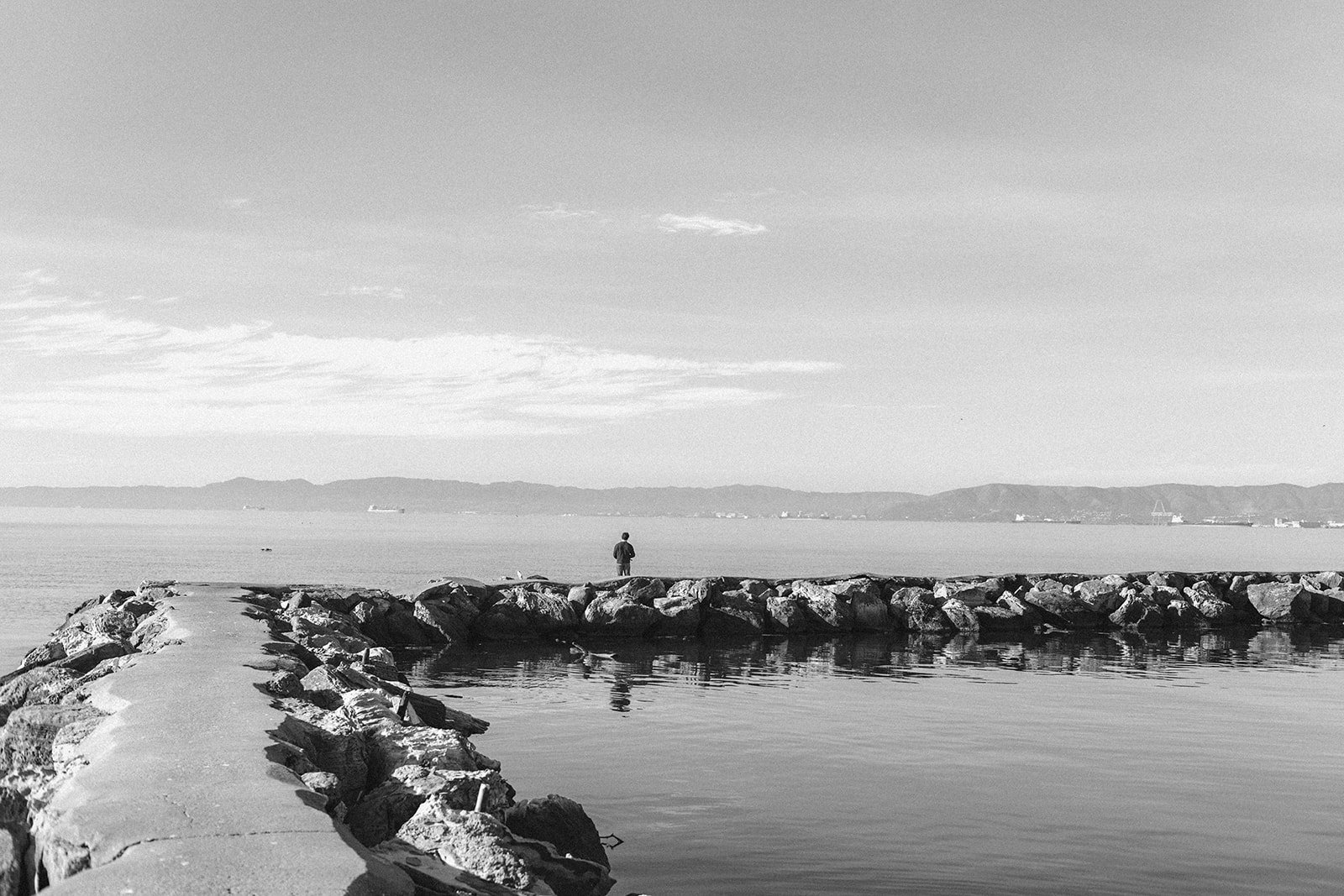 Black and white photo of a senior fishing from the Alameda Rock Wall with the San Francisco Bay in the distance.