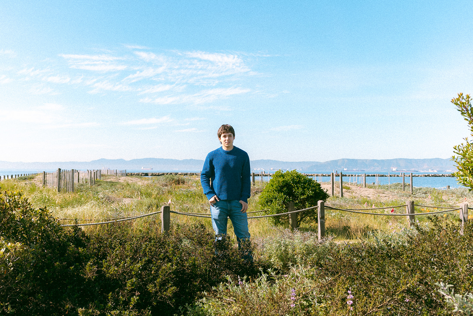 Senior standing among coastal greenery and spring wildflowers near the Alameda waterfront.
