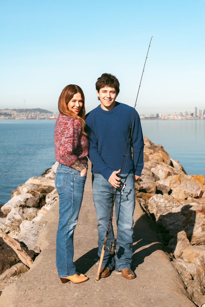 Senior posing with his mom and a fishing rod at the Alameda Rock Wall with the San Francisco skyline behind them.