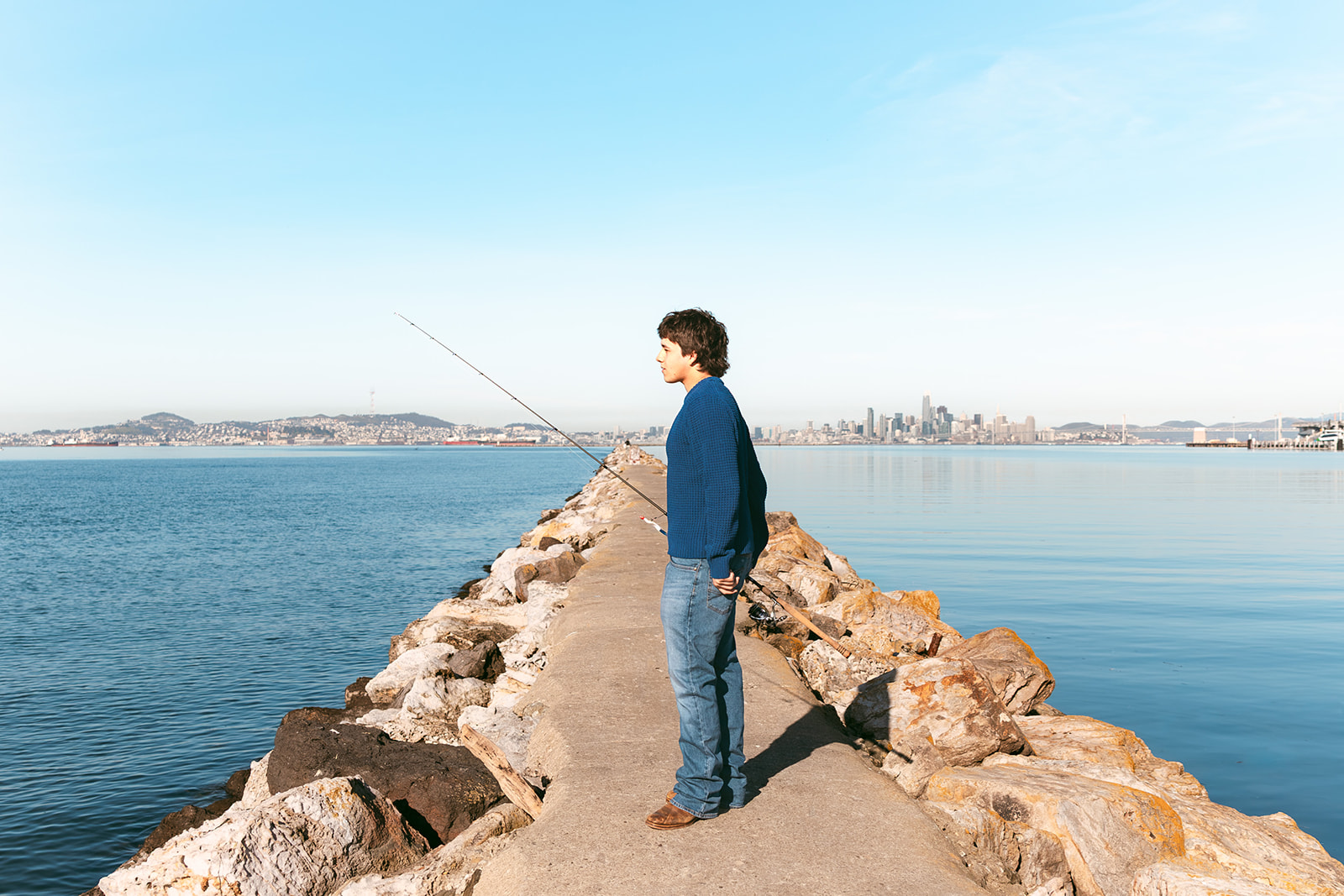 Senior holding a fishing rod on the Alameda Rock Wall with views of the San Francisco skyline across the Bay.