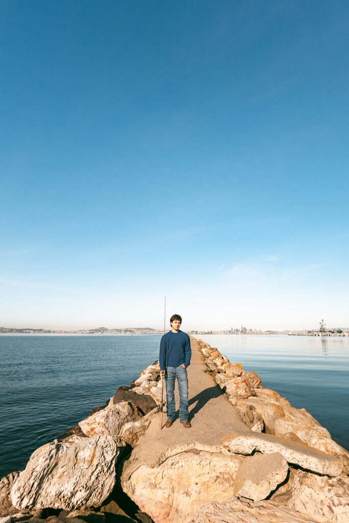 Senior standing with a fishing rod on the Alameda Rock Wall with the Bay and San Francisco skyline in the distance.