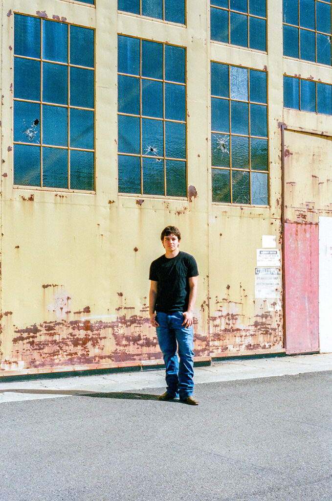 Film senior portrait standing in front of a weathered hangar with large industrial windows in Alameda.