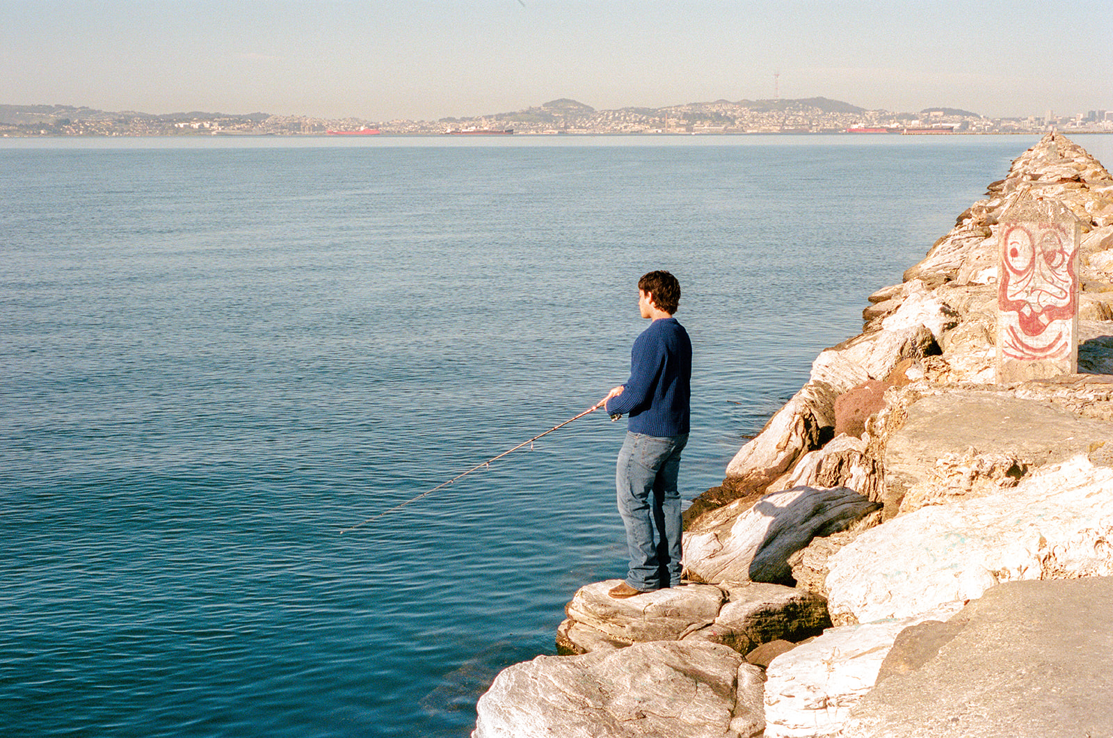 Film photo of a senior fishing from the rocks along the Alameda waterfront with the Bay behind him.