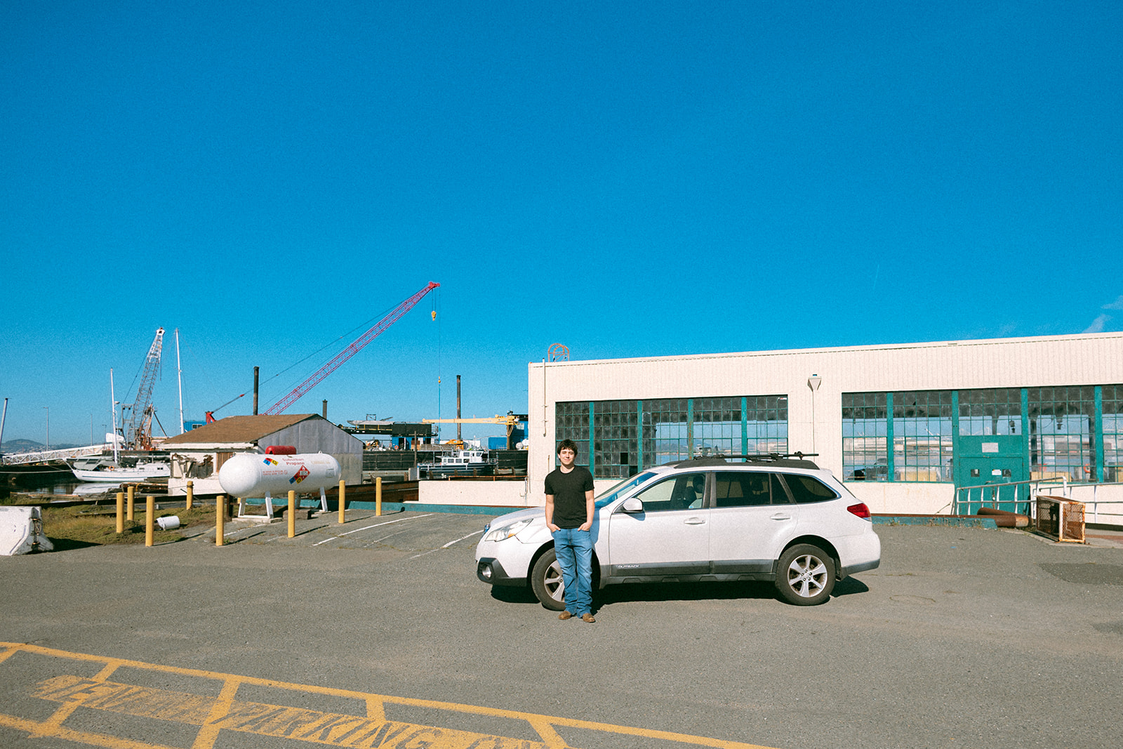 Senior standing beside his car near the former Naval Air Station in Alameda with industrial buildings and cranes behind him.