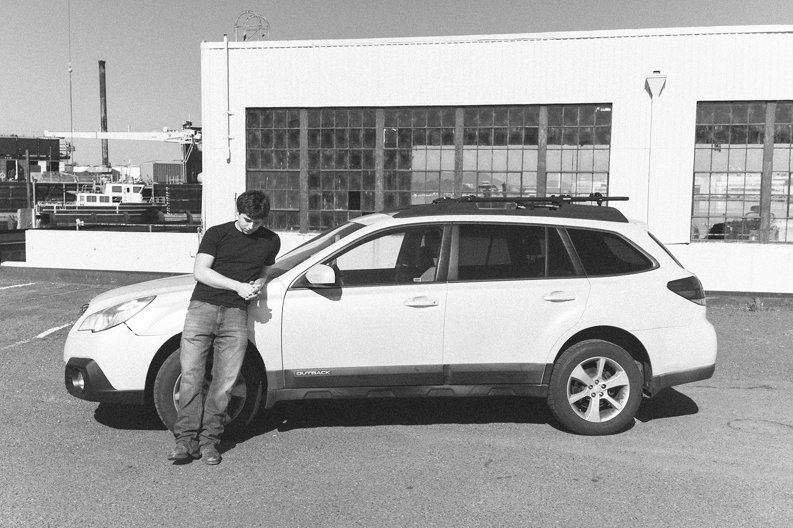 Black and white senior portrait leaning against his car outside an industrial warehouse in Alameda.
