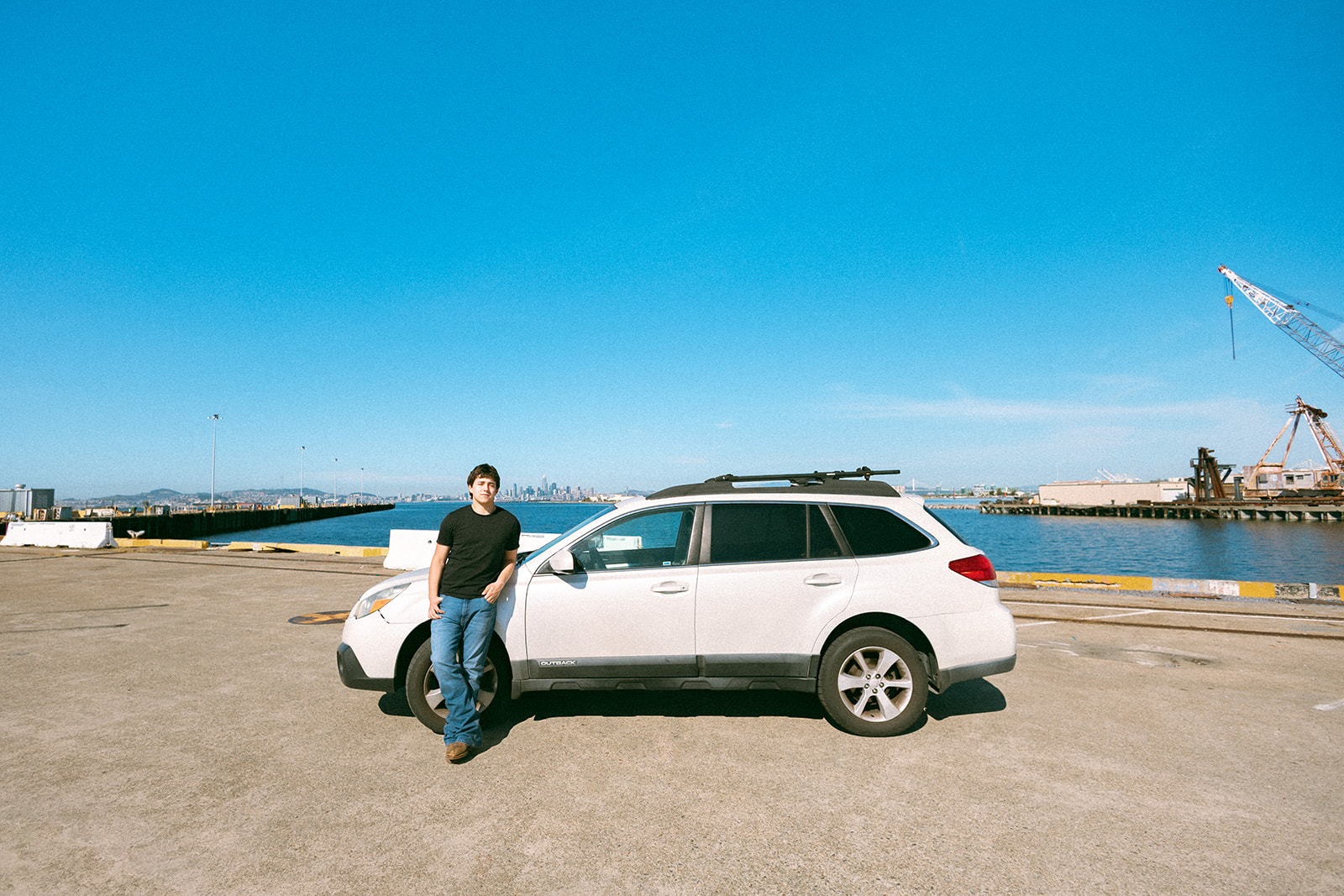Senior posing beside his car along the Alameda waterfront with cranes and the San Francisco skyline in the distance.