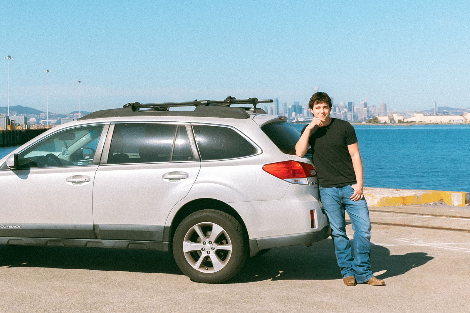 Senior leaning against his car with the San Francisco skyline visible across the Bay from Alameda.