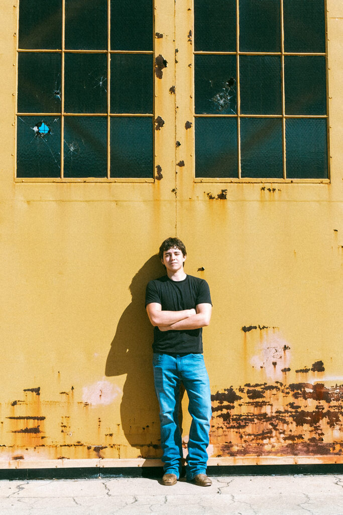 Senior portrait against a weathered yellow hangar with broken windows at the former Alameda Naval Air Station.