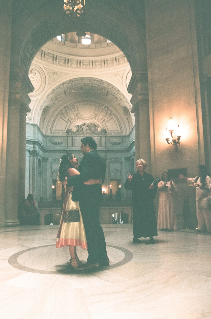 Bride and groom hugging during their San Francisco City Hall wedding as family watches nearby.
