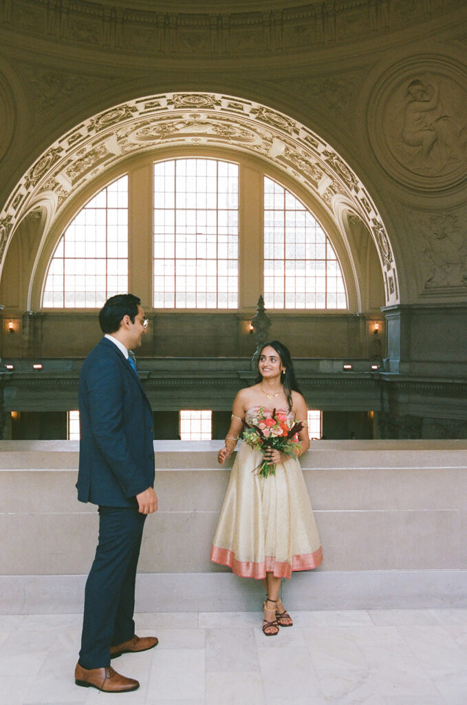 Bride and groom posing beneath a large arched window inside San Francisco City Hall.