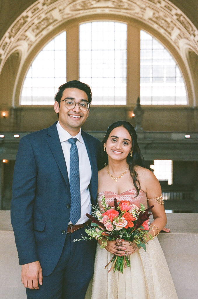 Bride and groom smiling beneath a large arched window inside San Francisco City Hall.