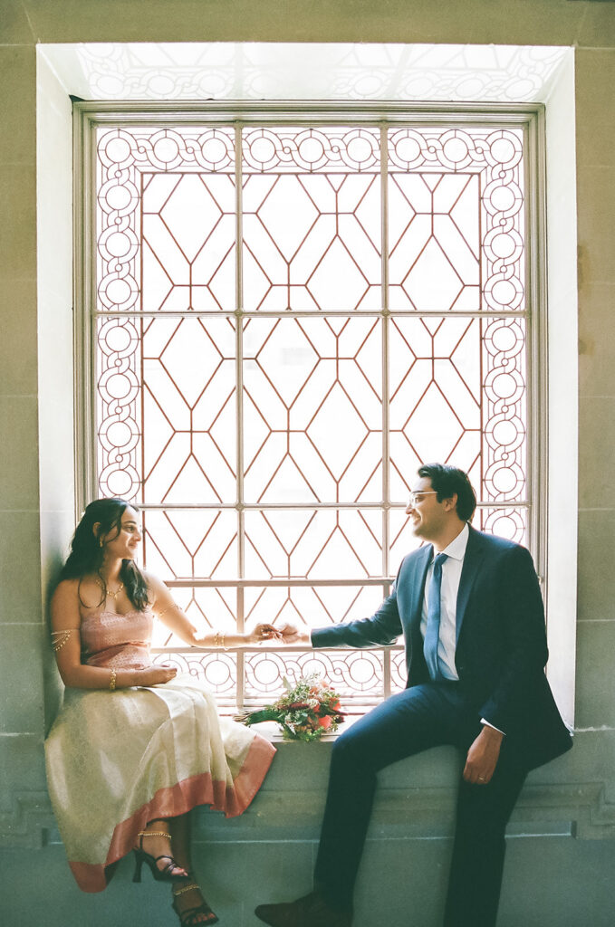 Newlyweds sitting by a decorative window and holding hands at San Francisco City Hall.