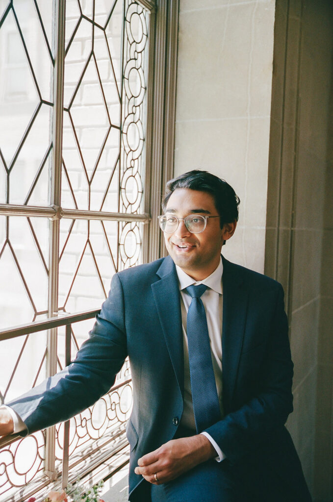 Groom sitting by a decorative window and holding hands with his bride at San Francisco City Hall.