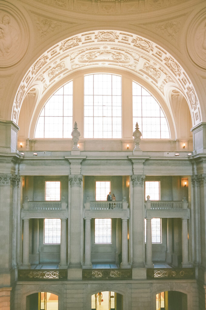 Wide film photograph of the newlyweds on an upper balcony inside San Francisco City Hall.