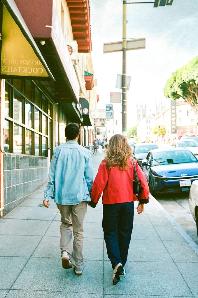 Couple walking together along a North Beach sidewalk lined with neighborhood storefronts.