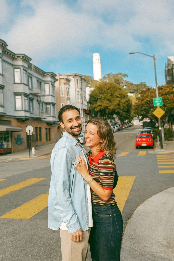 Engaged couple laughing together on a San Francisco street with Coit Tower in the background.